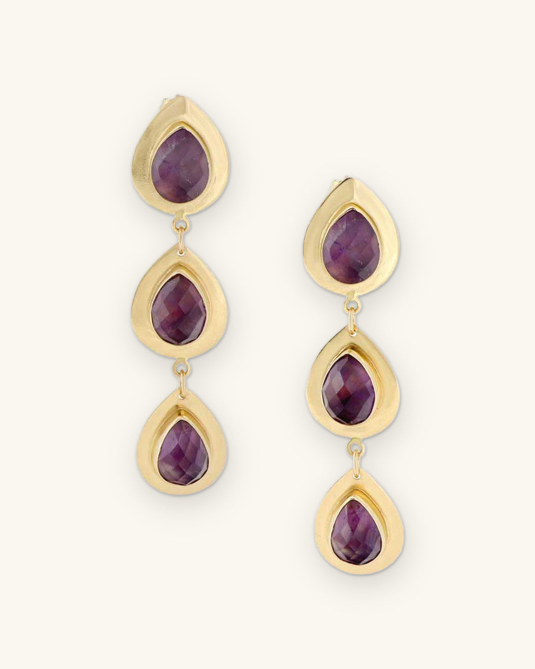Triple amethyst teardrop drop earrings with faceted stones bezel-set in brass, made in India, front view, shown on a sand background