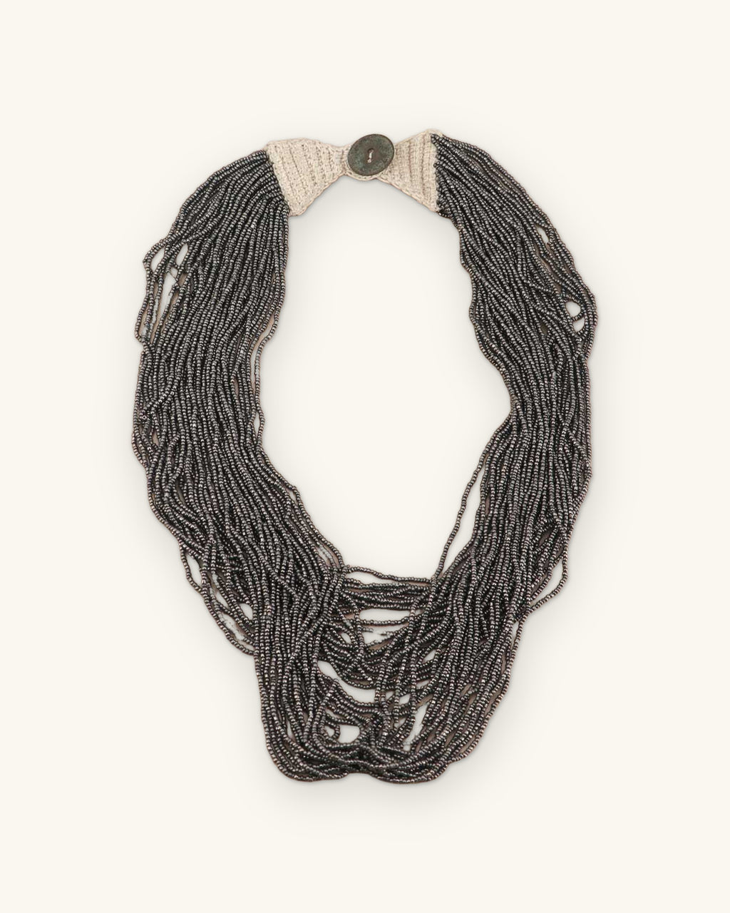 Antique gray multi-strand seed bead necklace laid flat with cream crochet ends and a button closure at top, shown on a sand background