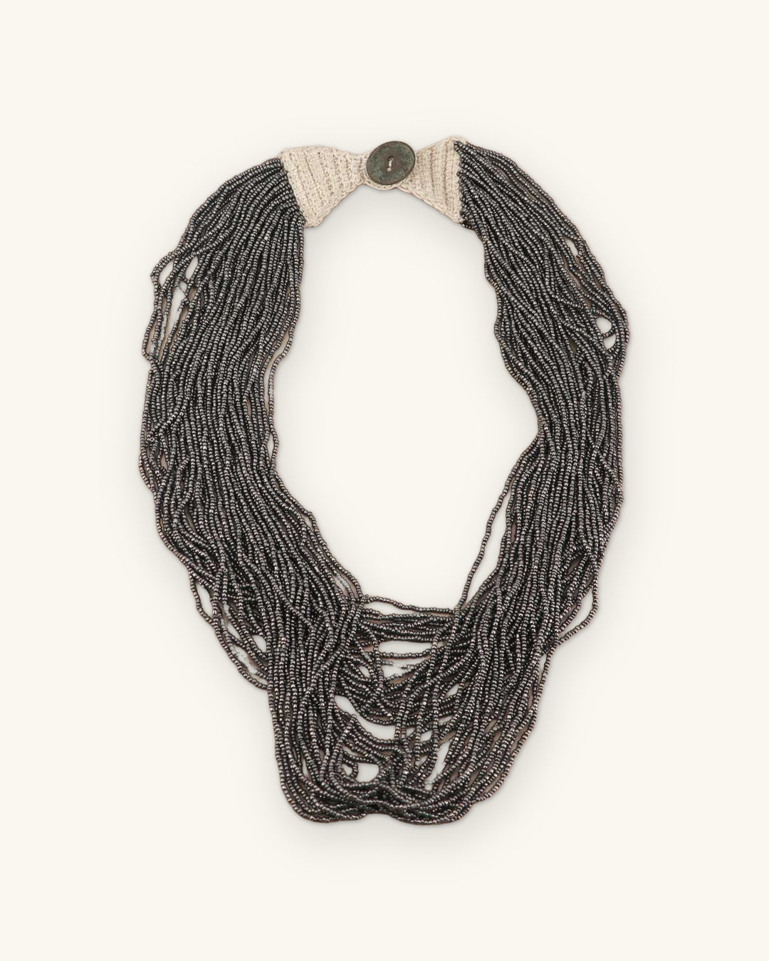 Antique gray multi-strand seed bead necklace laid flat with cream crochet ends and a button closure at top, shown on a sand background