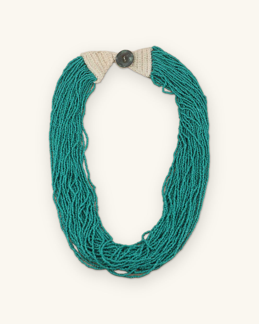 Multi-strand turquoise seed bead necklace laid flat with cream crocheted cotton ends and a button closure, shown on a sand background