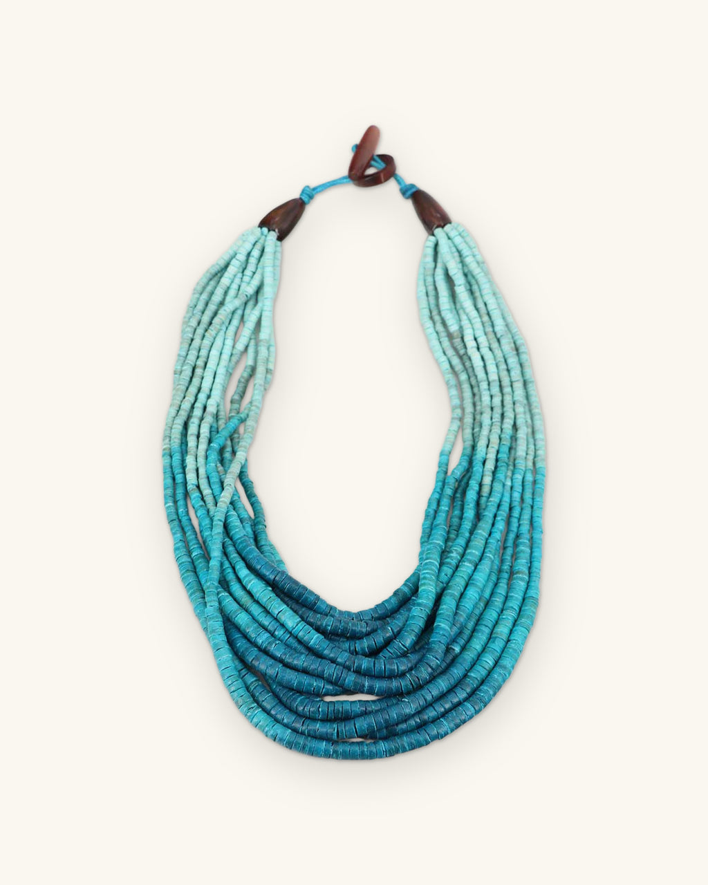 Multi-strand coconut bead bib necklace fading from pale aqua to deep teal, with wooden toggle closure, shown on a sand background