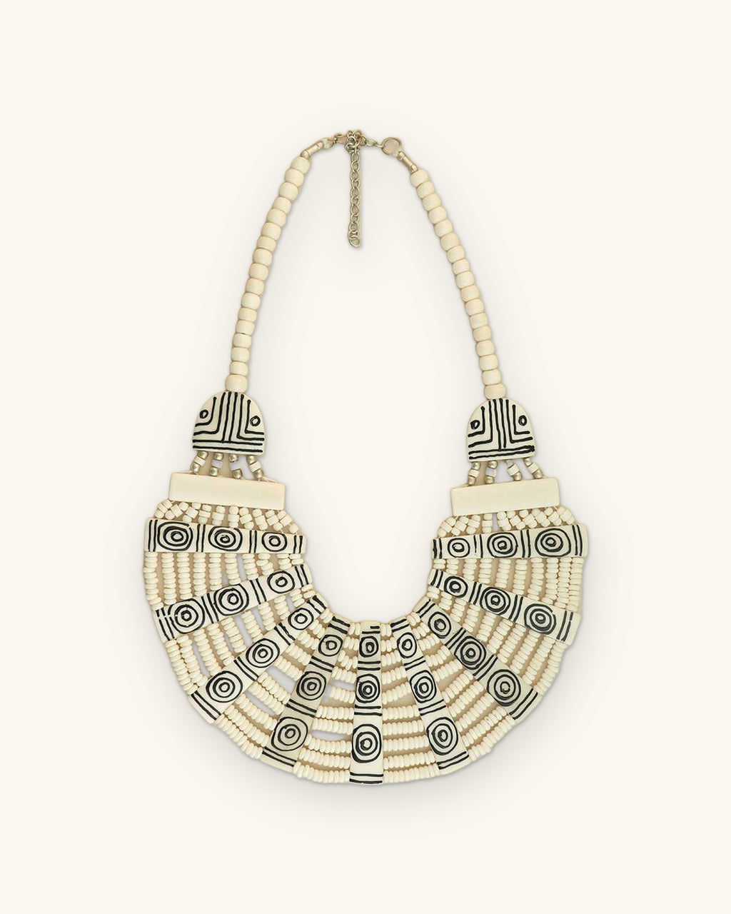 Graphic carved bone bib necklace laid flat, black spiral and line motifs on cream bone with bead strands, shown on a sand background
