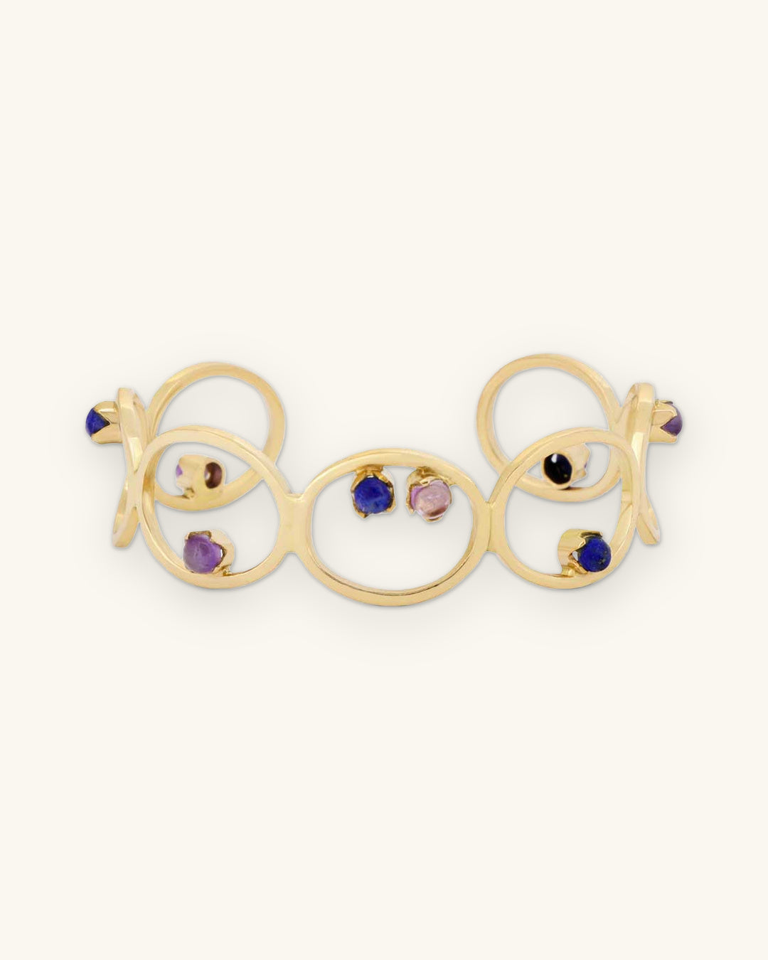 Interlocking circle brass cuff with prong-set amethyst and lapis lazuli cabochons, hand-fabricated in India, shown on a sand background