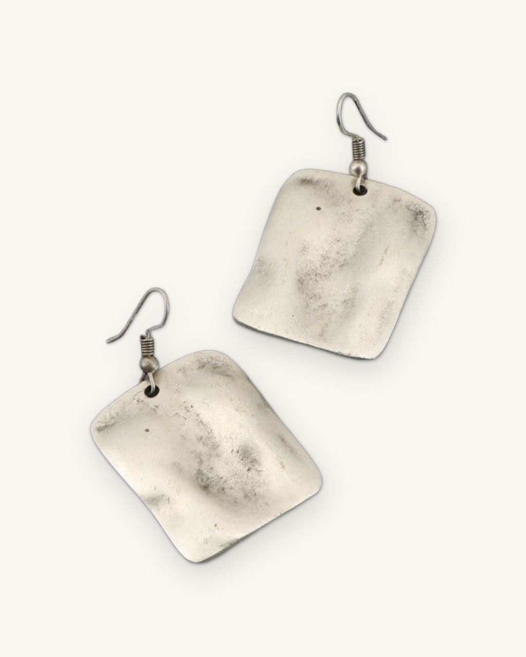 Pair of brushed silver-tone rectangle earrings with a rippled wave surface on fish-hook ear wires, shown on a sand background