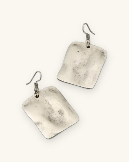 Pair of brushed silver-tone rectangle earrings with a rippled wave surface on fish-hook ear wires, shown on a sand background