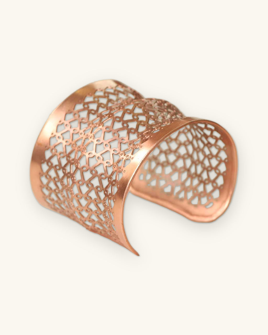 Pierced copper lattice wide cuff bracelet with a geometric cutout pattern, handmade in India, shown on a sand background