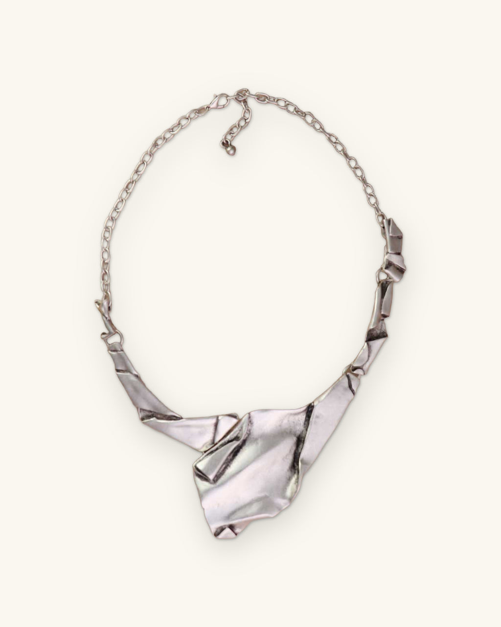 Full view of the crumpled-paper style silver-tone bib necklace with folded links and cable chain, shown on a sand background