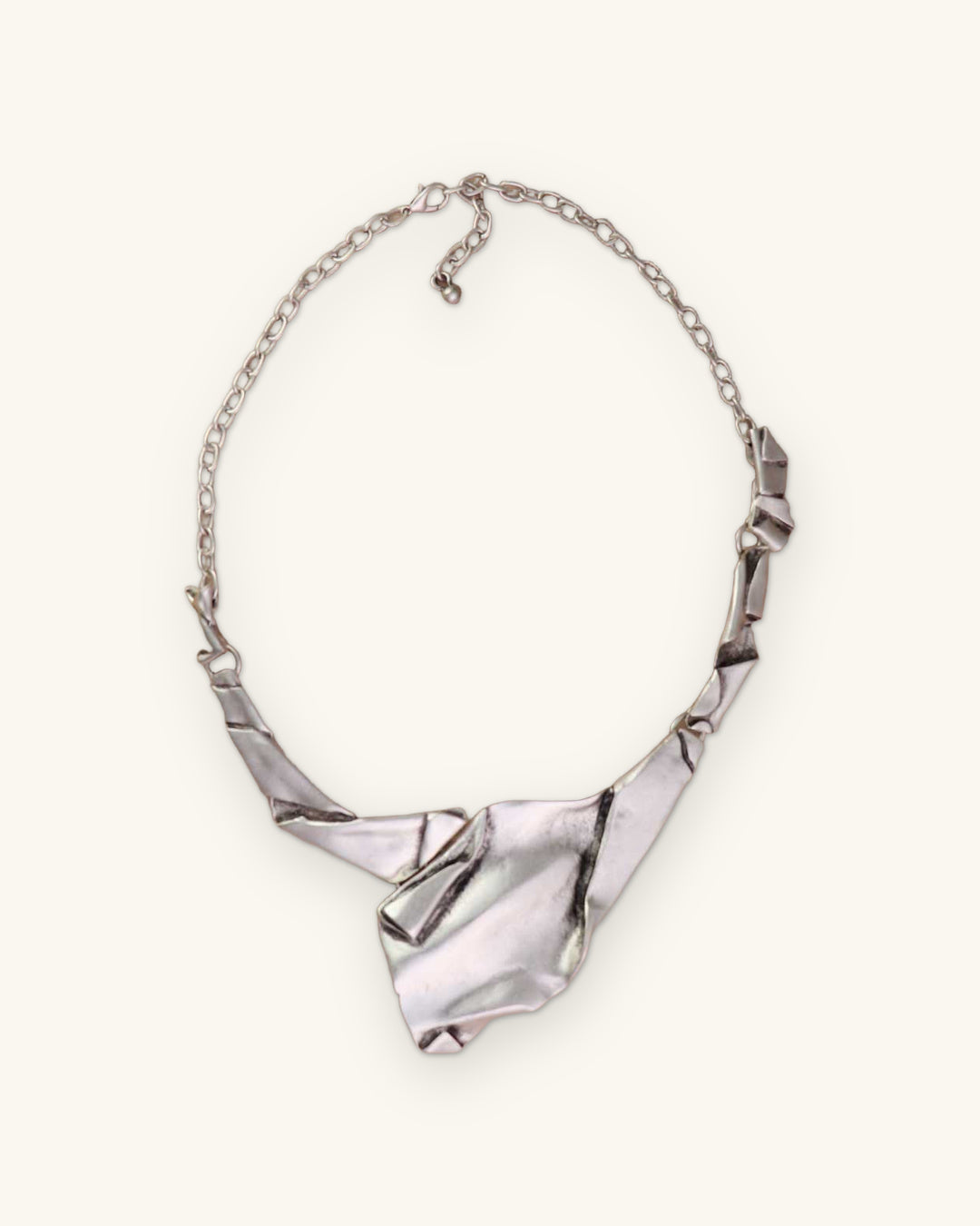 Full view of the crumpled-paper style silver-tone bib necklace with folded links and cable chain, shown on a sand background