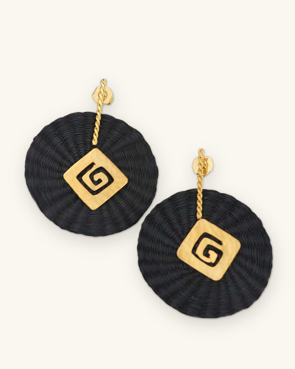 Woven black iraca palm disc earrings with gold-plated spiral charms, handwoven in Colombia with hammered metalwork, shown on a sand background