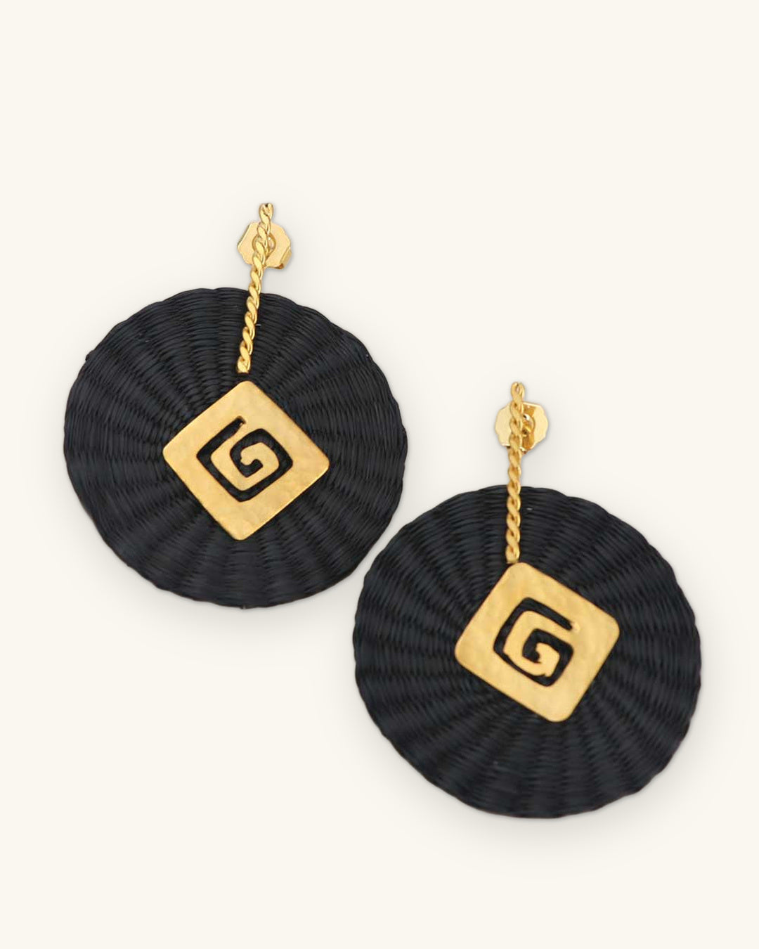 Woven black iraca palm disc earrings with gold-plated spiral charms, handwoven in Colombia with hammered metalwork, shown on a sand background
