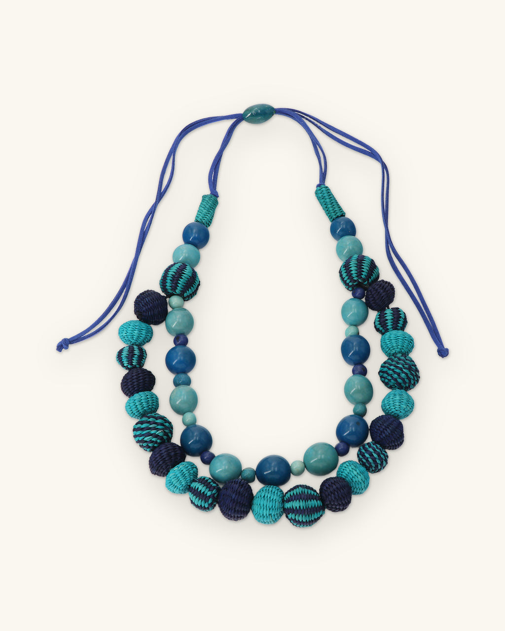 Teal and navy woven bead double-strand necklace with adjustable blue cord and sliding bead, shown on a sand background