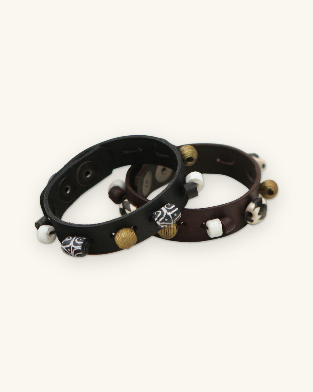 Beaded leather snap bracelets in black and brown with painted, brass and white disc beads, made in Ghana, shown on a sand background