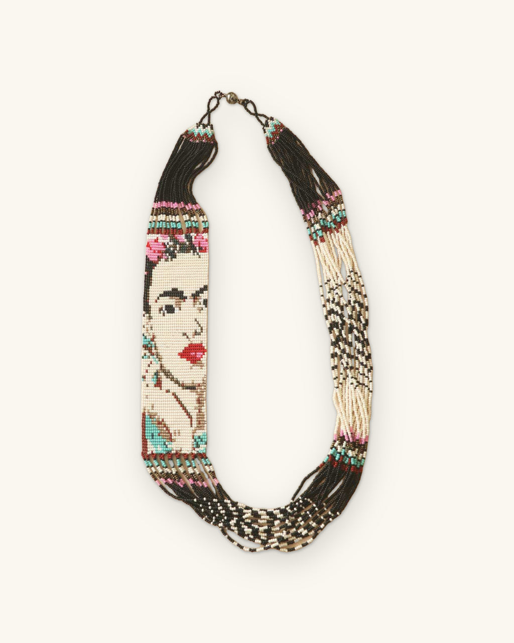 Frida Kahlo seed bead portrait necklace laid flat, cream strands with black and white patterned fringe, shown on a sand background