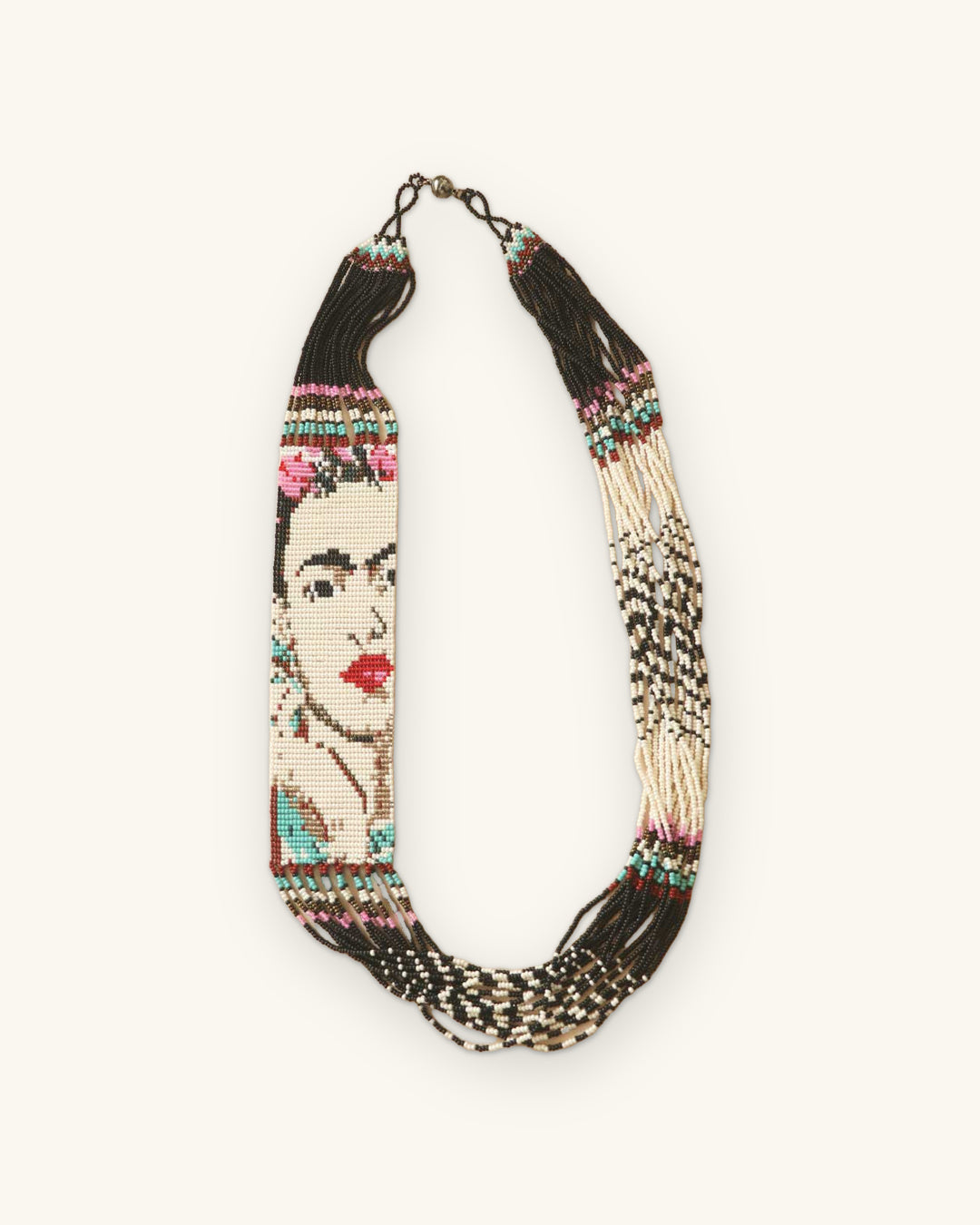 Frida Kahlo seed bead portrait necklace laid flat, cream strands with black and white patterned fringe, shown on a sand background