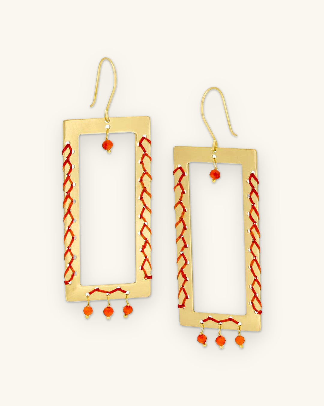 Brass rectangle frame drop earrings laced with orange thread and carnelian bead drops, handmade in India, shown on a sand background