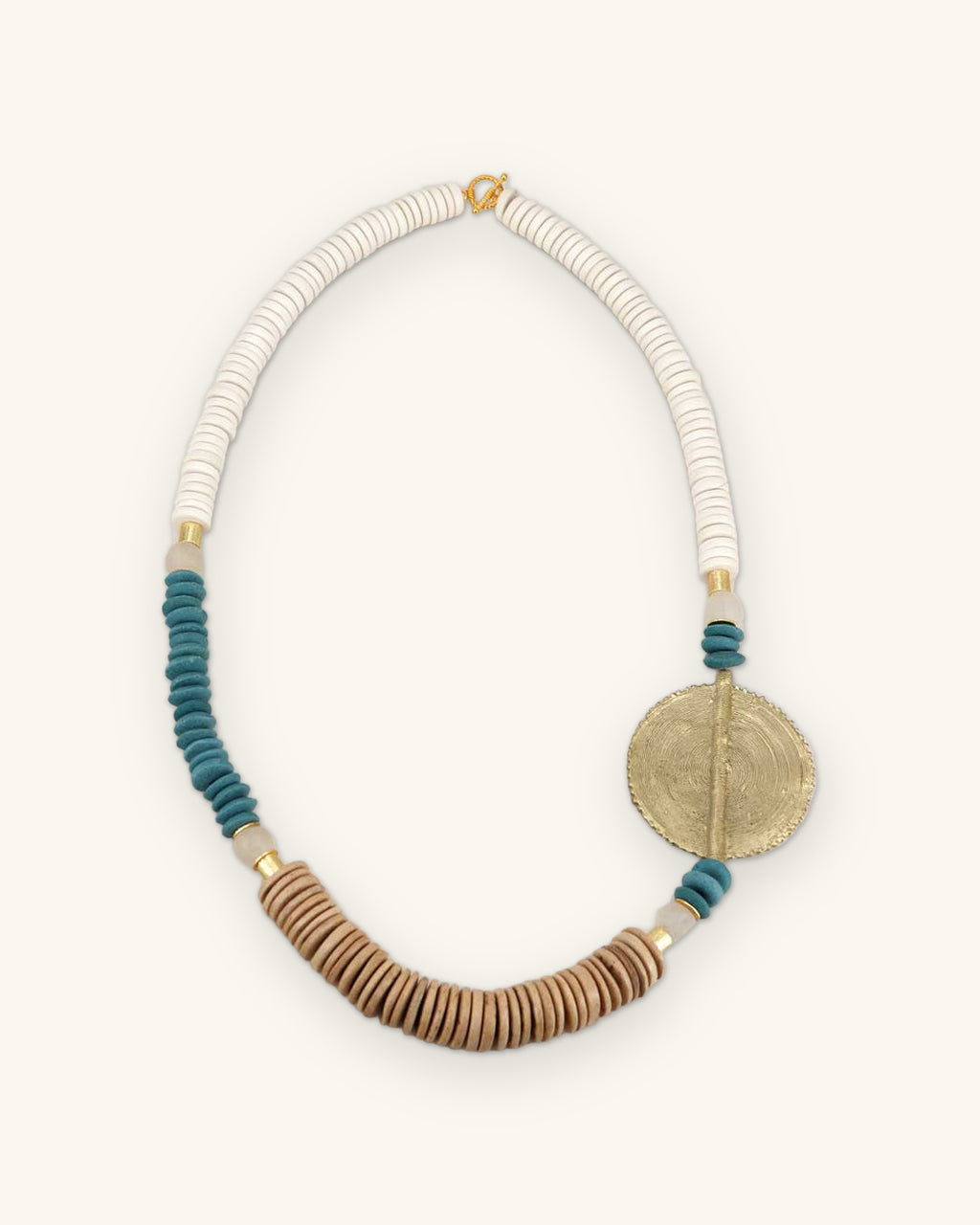 White bone, teal and tan disc bead necklace with round engraved brass medallion, shown on a sand background