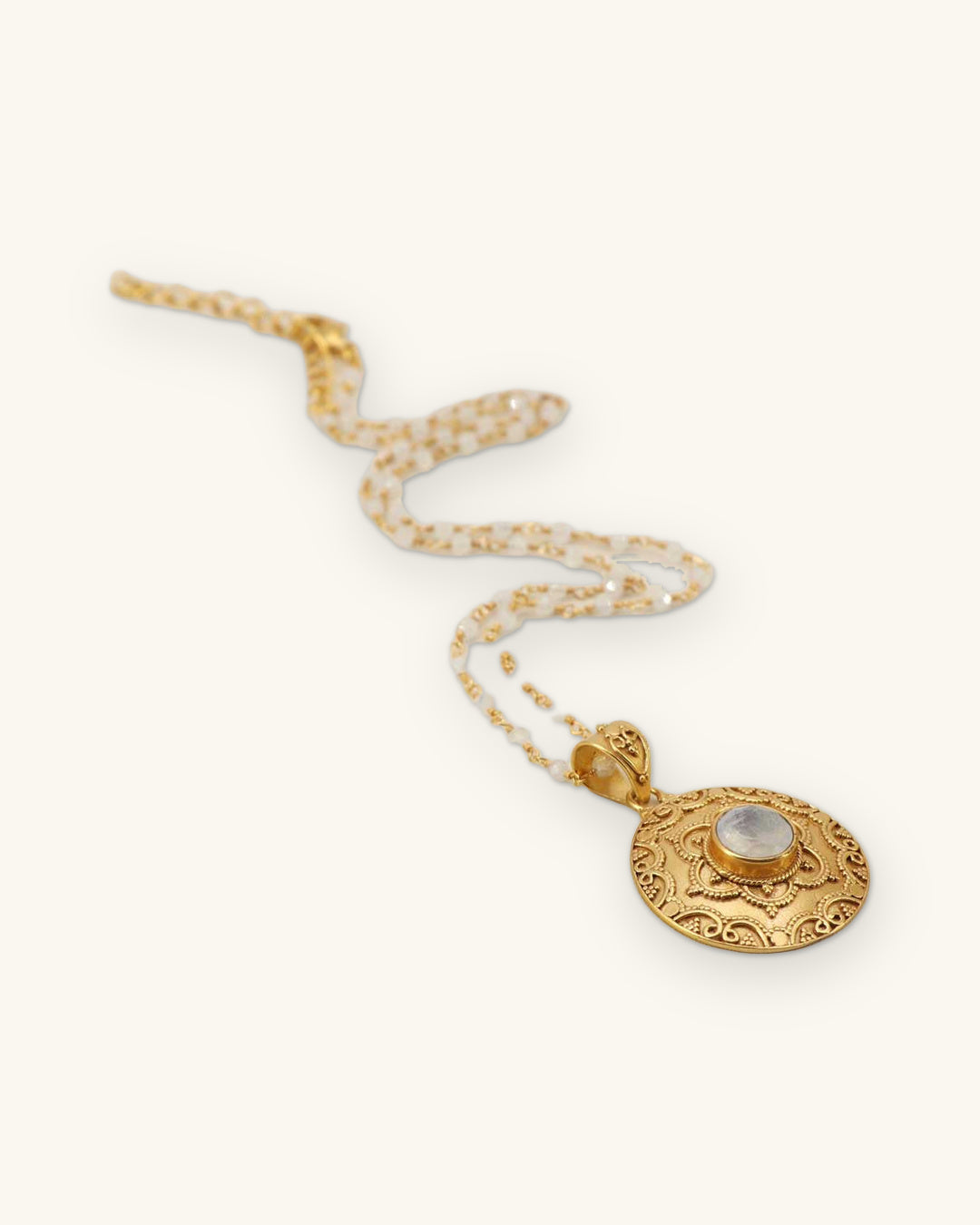 Engraved gold-plated brass floral medallion necklace with a rainbow moonstone center on beaded chain, made in India, shown on a sand background