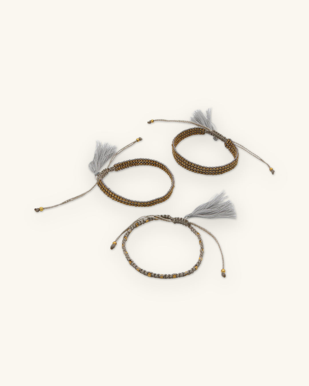 Set of three beaded cord bracelets in Gray with gold-tone beads and adjustable tasseled ties, shown on a sand background