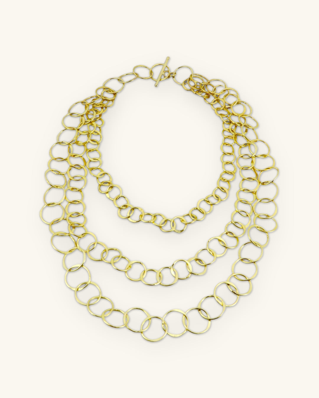 Graduated three-strand brass chain necklace laid flat with toggle clasp, shown on a sand background
