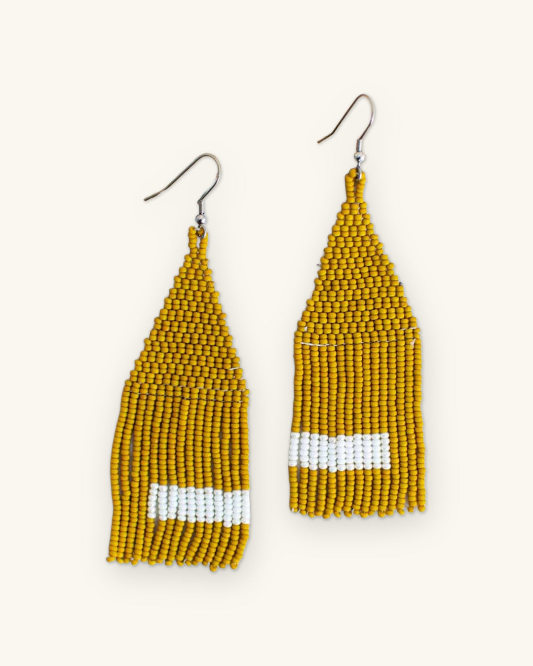 Pair of mustard seed bead fringe earrings with white bands on a ceramic plate, shown on a sand background