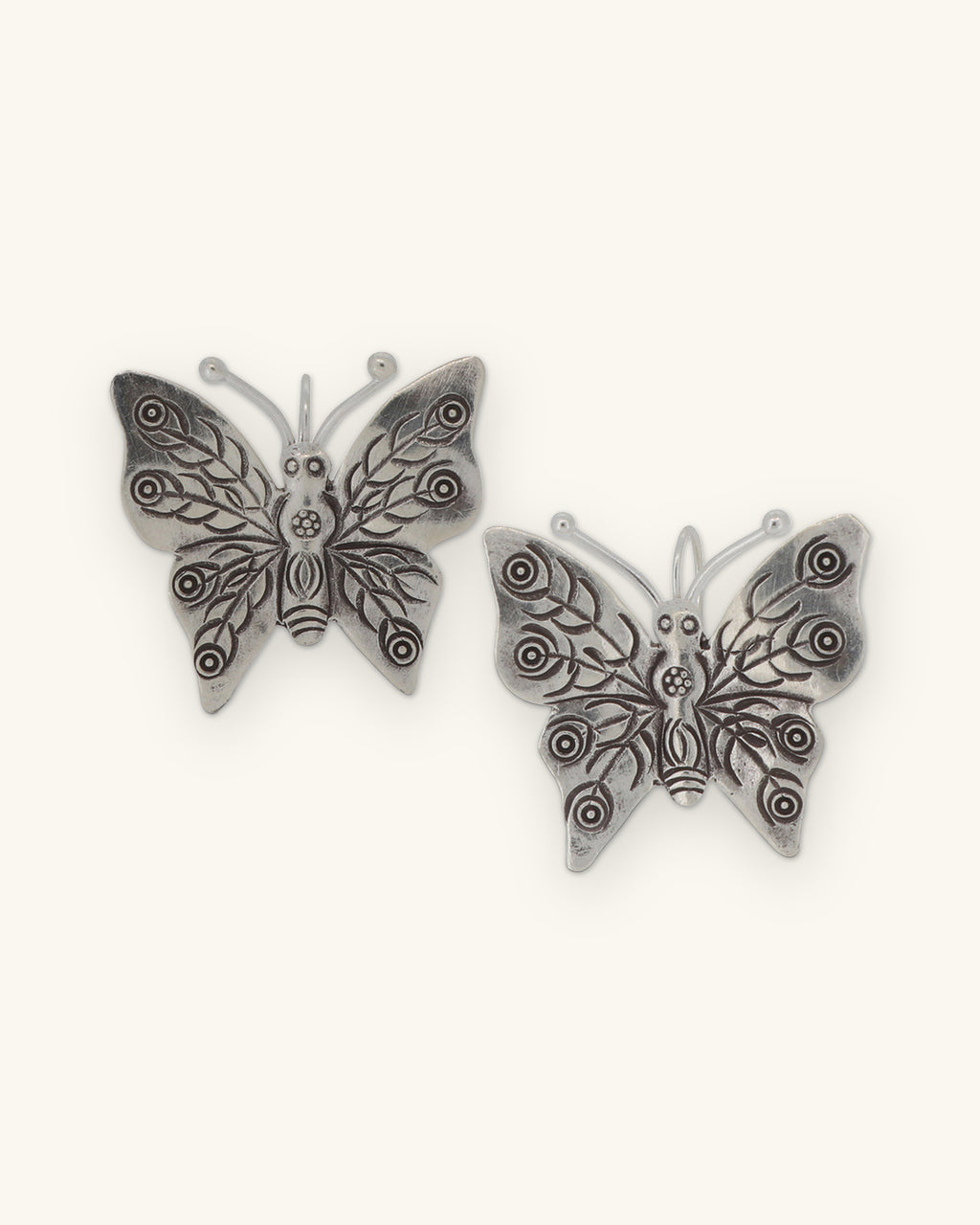 Pair of silver butterfly earrings with stamped swirl and leaf wing patterns, shown on a sand background