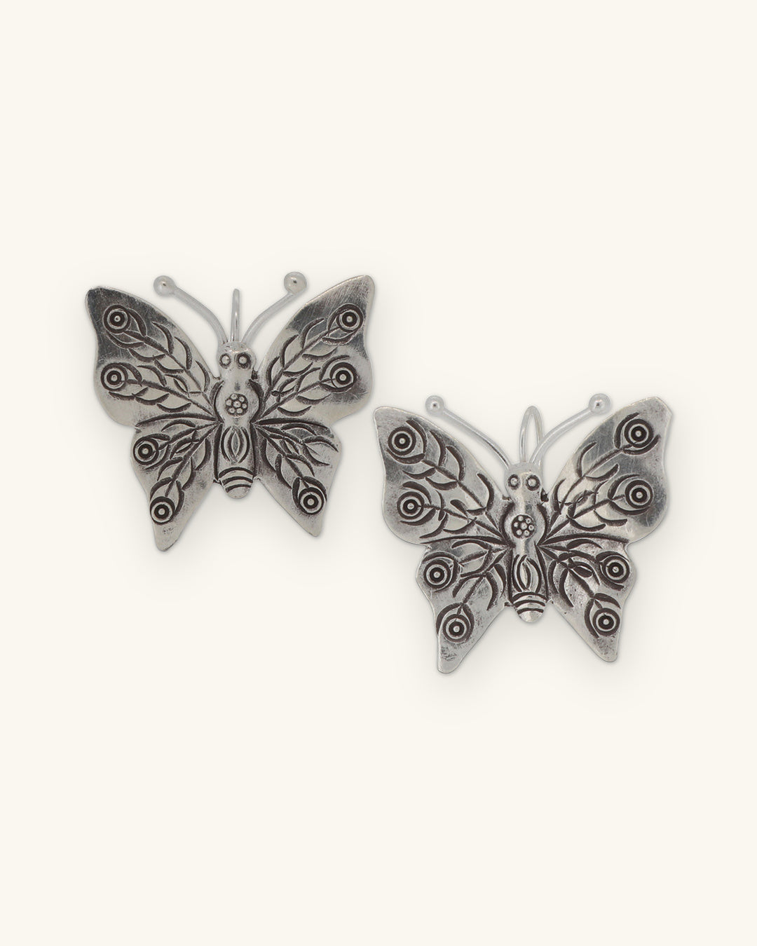 Pair of silver butterfly earrings with stamped swirl and leaf wing patterns, shown on a sand background