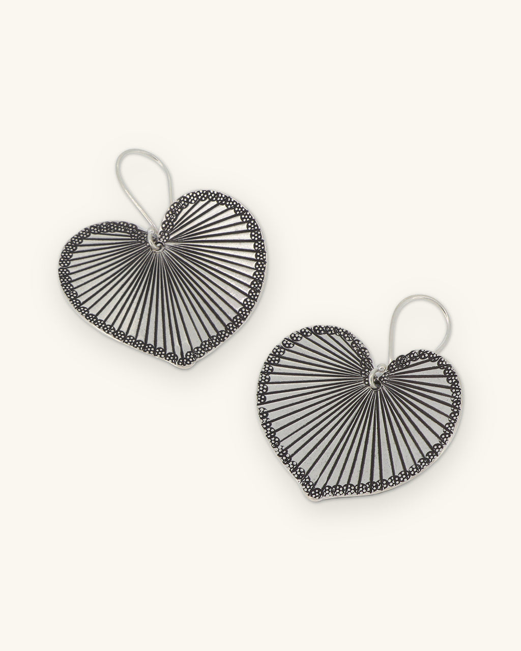 Pair of silver heart-leaf drop earrings with radiating engraved veins and beaded rims, shown on a sand background