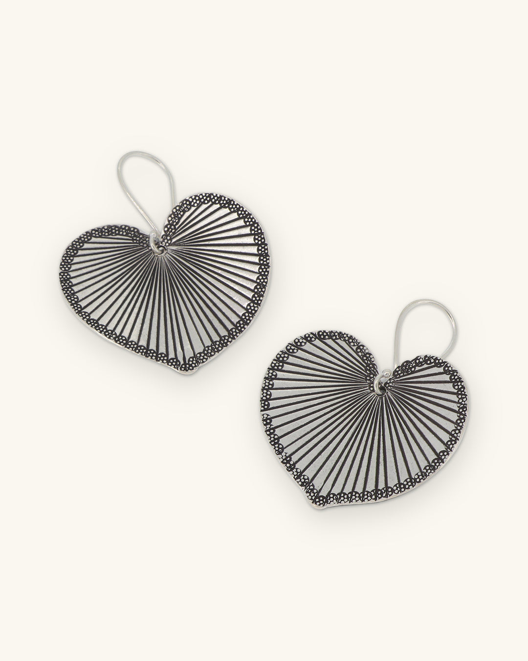 Pair of silver heart-leaf drop earrings with radiating engraved veins and beaded rims, shown on a sand background