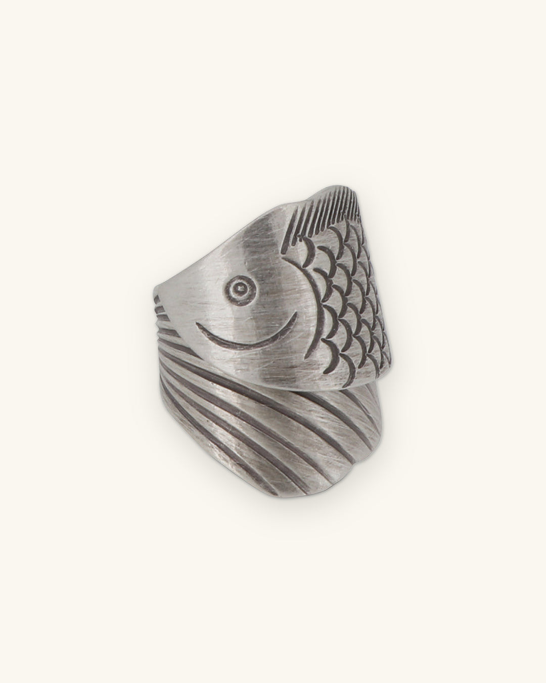Side view of the silver fish-wrap ring showing the engraved eye, smile and scales, shown on a sand background