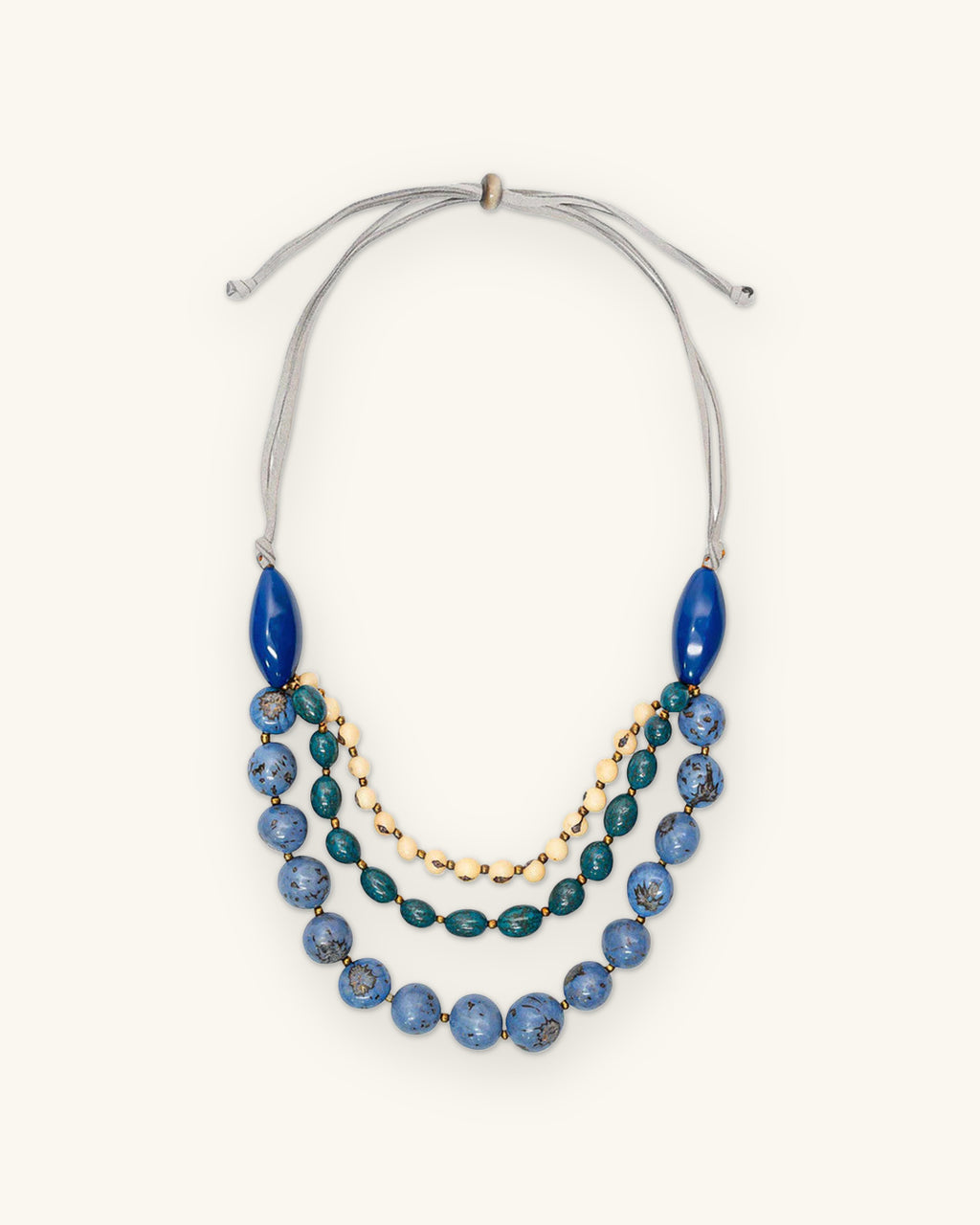 Three-strand blue tagua nut and seed bead necklace with cream bead strand and adjustable cord, handmade in Ecuador, shown on a sand background