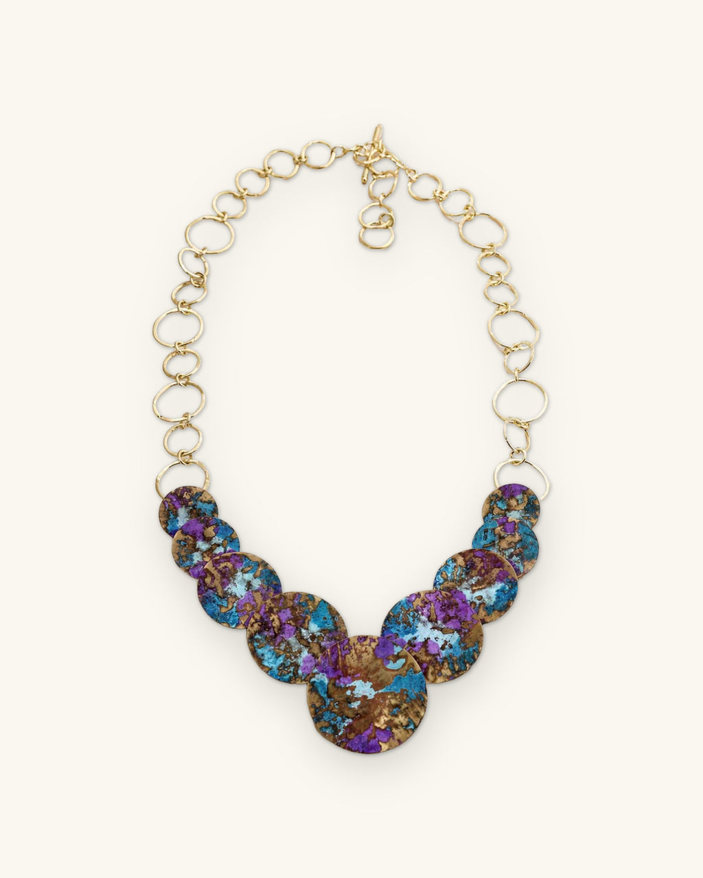 Brass disc bib necklace with violet and turquoise patina on a gold-tone ring chain, hand-forged in India, shown on a sand background