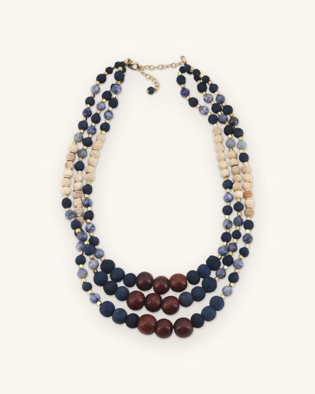 Graduated three-strand necklace with navy fabric beads, sodalite, cream wood beads and a mahogany bead center, shown on a sand background