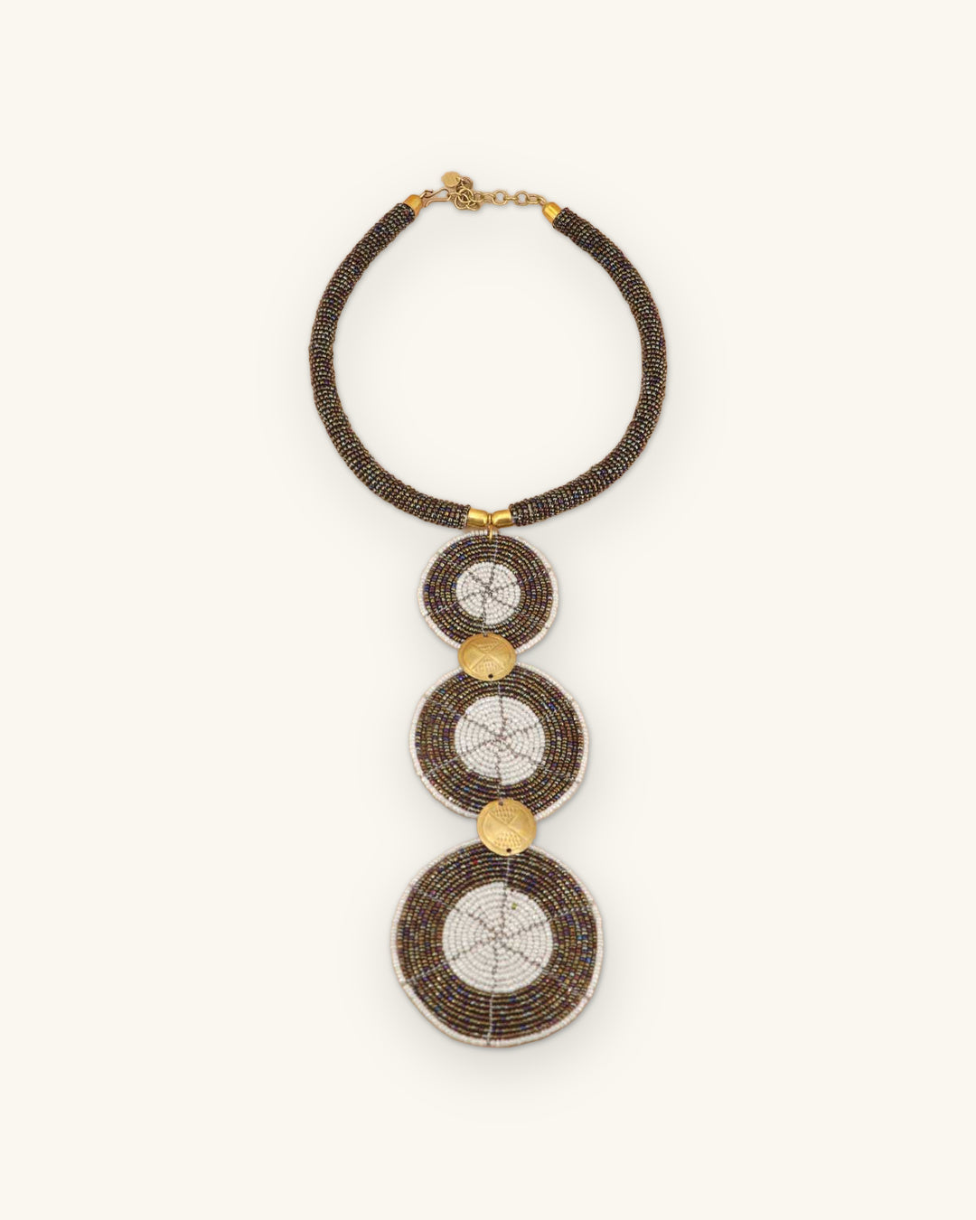 Brown glass bead collar necklace laid flat with three white-centered bead discs linked by brass coins, shown on a sand background