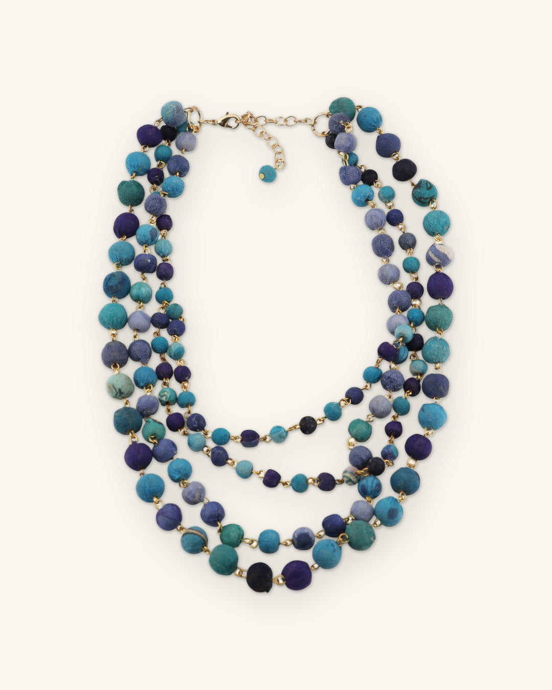 Four-strand blue fabric bead necklace laid flat with lobster clasp and extender chain, shown on a sand background