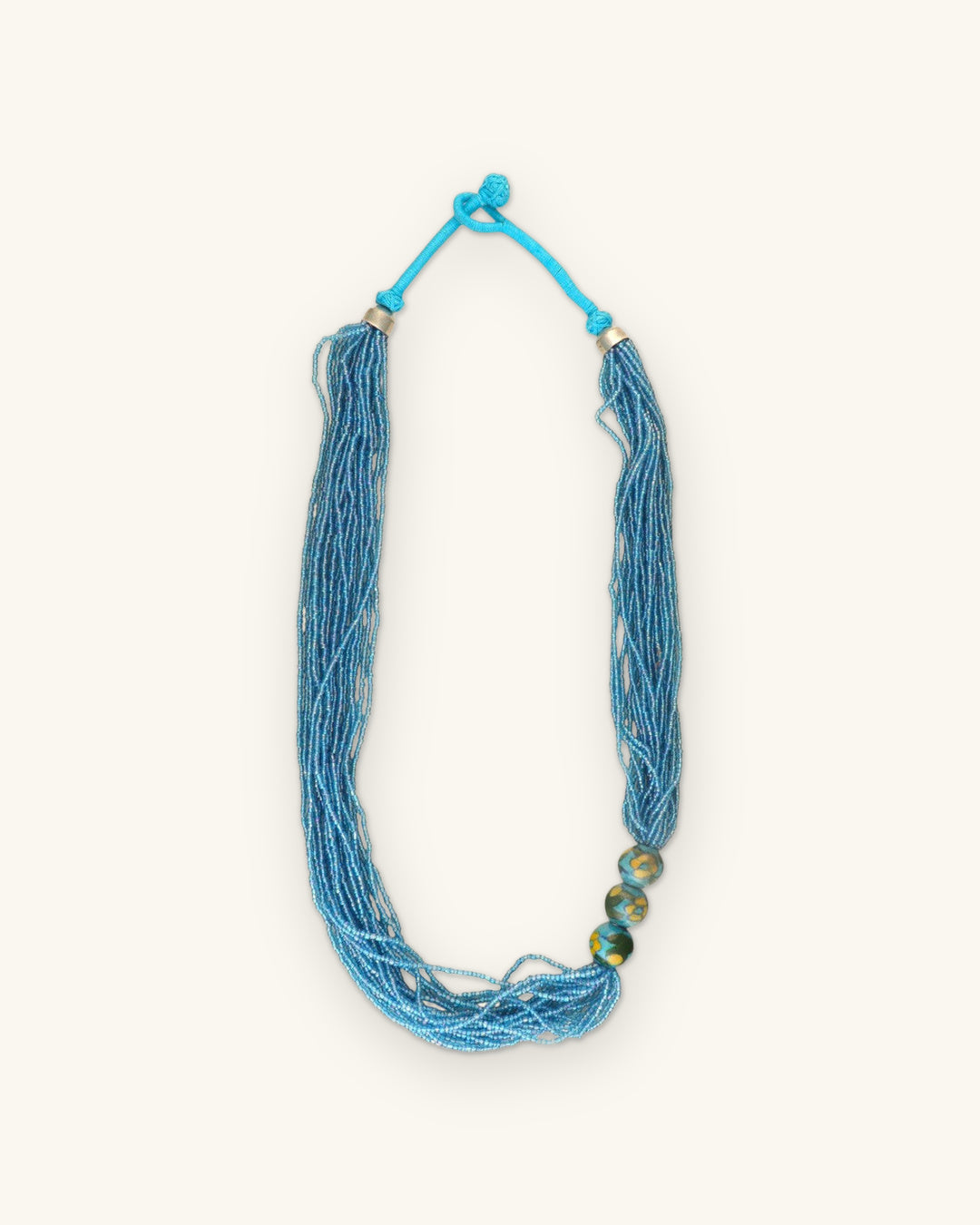 Full view of the aqua seed bead rope necklace with painted ceramic trio and knotted cord closure, shown on a sand background