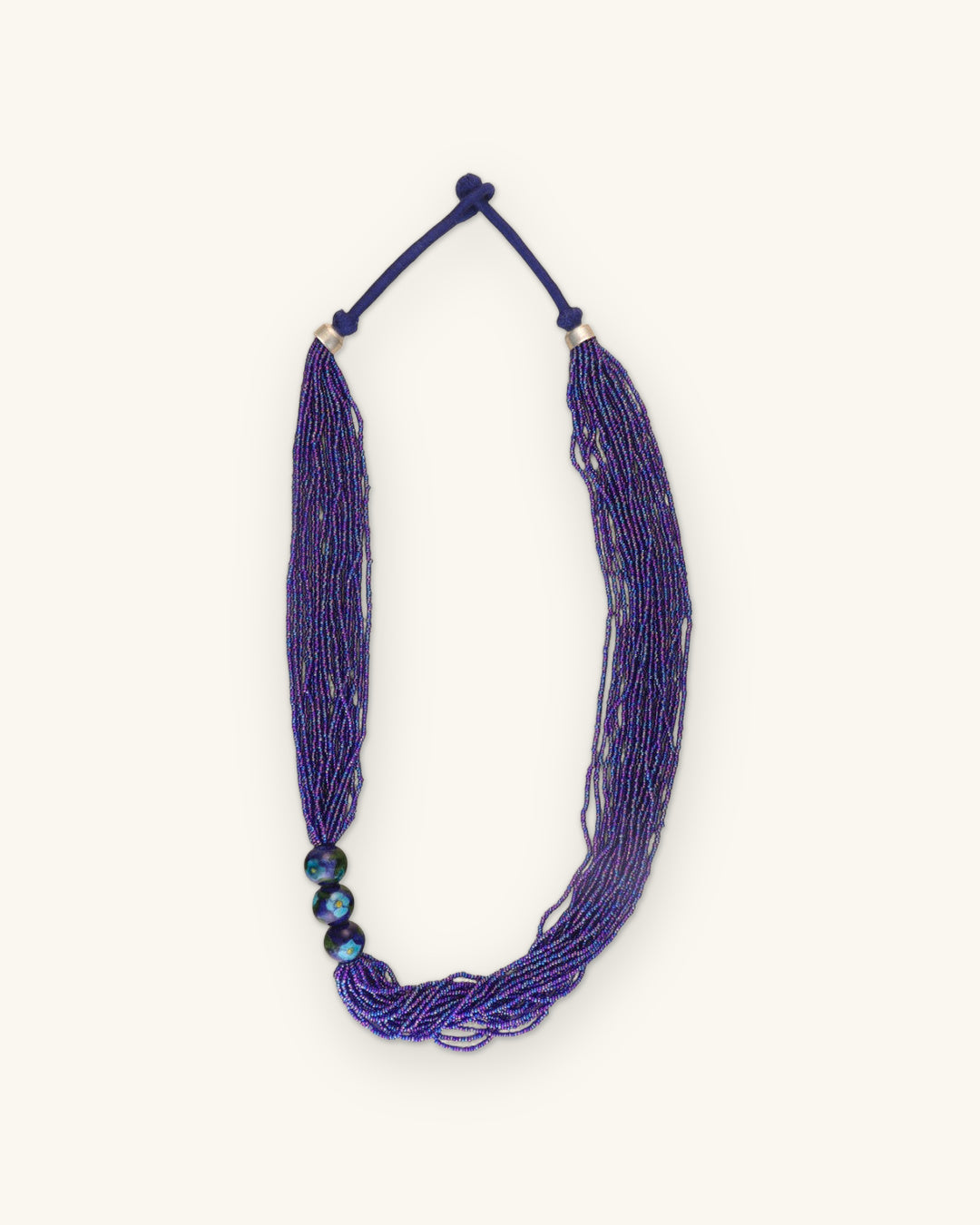 Full view of the violet multi-strand seed bead necklace with knotted cord closure and silver-tone caps, shown on a sand background