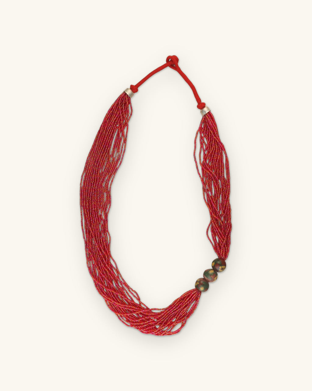 Full view of the red seed bead necklace with knotted red cord closure and silver-tone caps, shown on a sand background