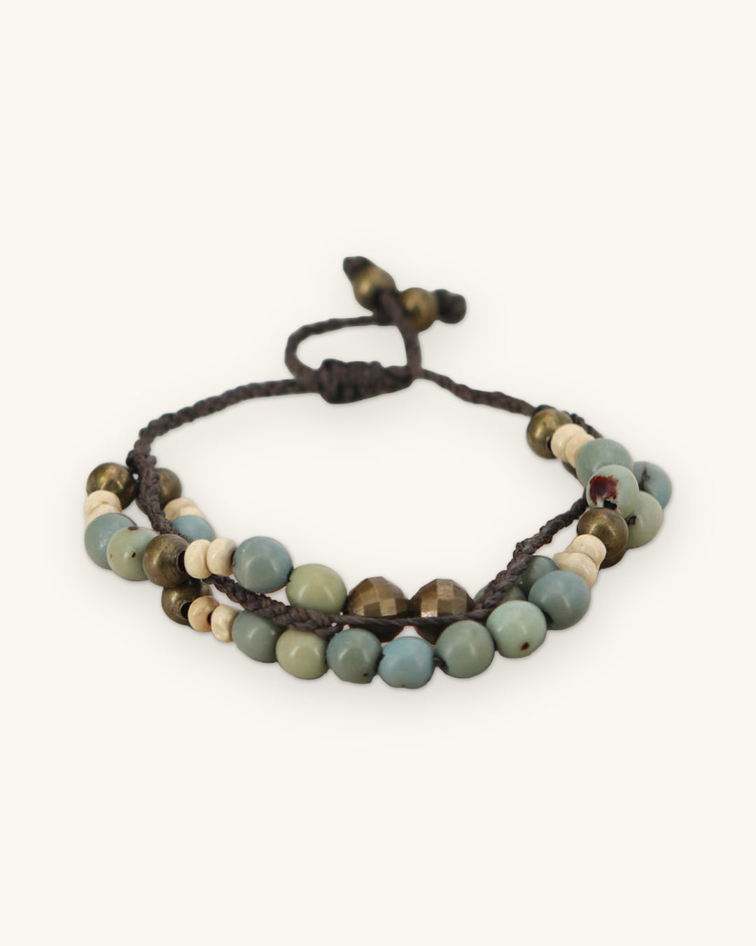 Two-strand blue-green seed bead bracelet with faceted brass beads on a beige surface, shown on a sand background