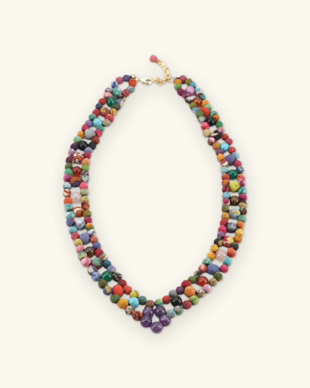 Full view of the multicolor textile and gemstone bead necklace with gold-tone clasp, shown on a sand background