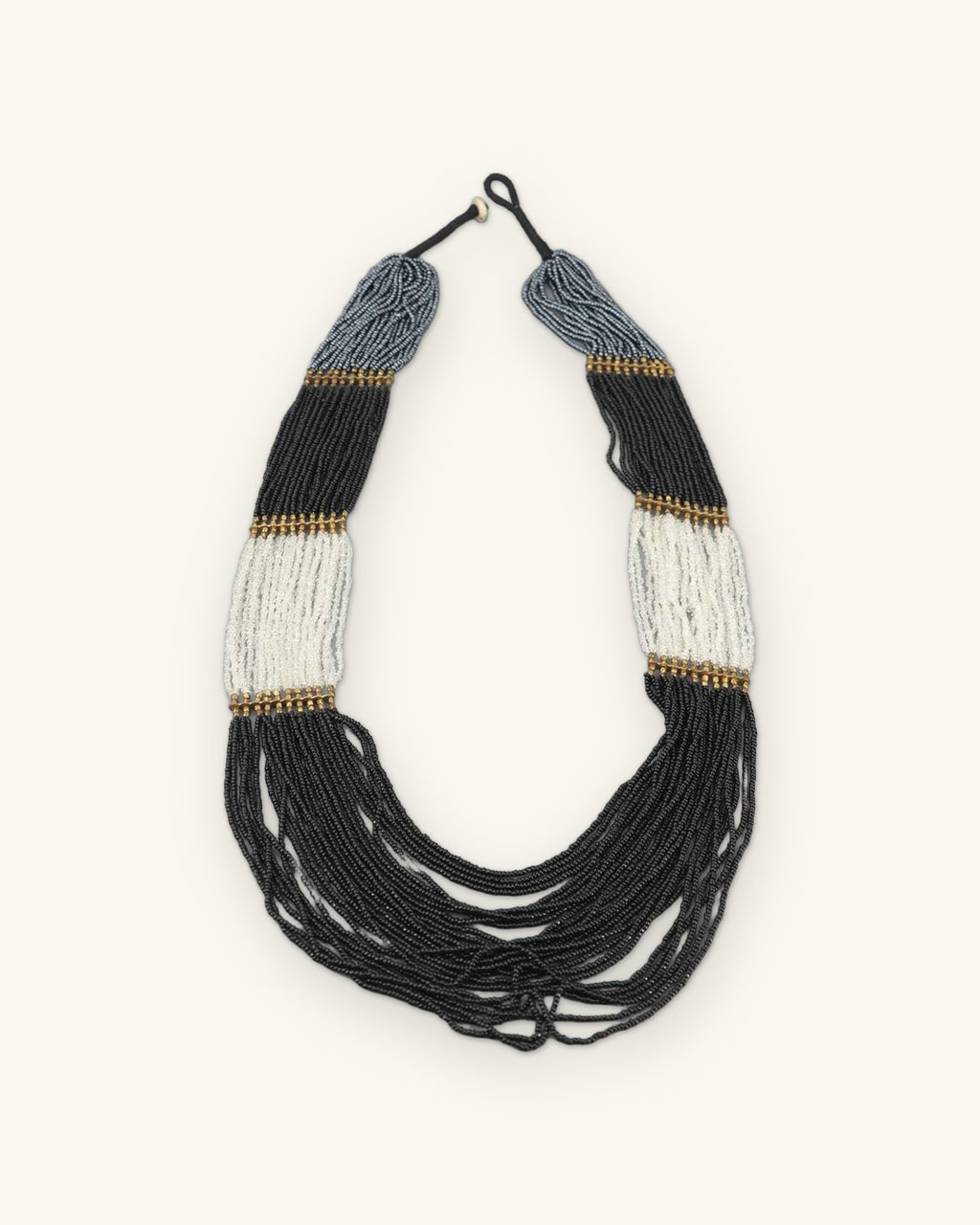 Full view of the black, gray and ivory seed bead necklace with brass-tone bands and knotted cord closure, shown on a sand background