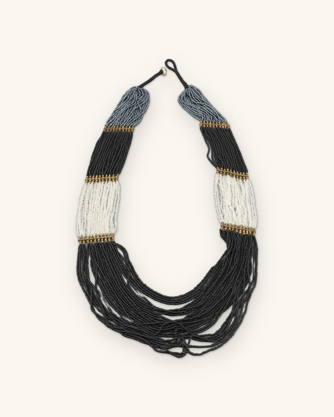 Full view of the black, gray and ivory seed bead necklace with brass-tone bands and knotted cord closure, shown on a sand background