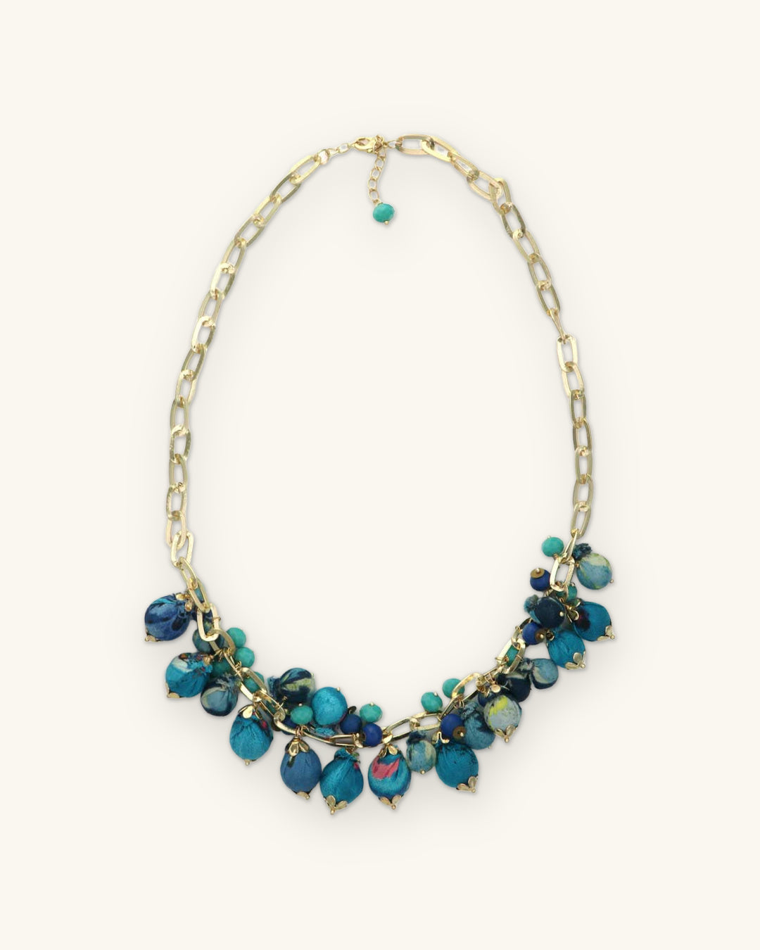 Full view of the teal and blue fabric bead cluster necklace on a gold-tone paperclip chain, shown on a sand background