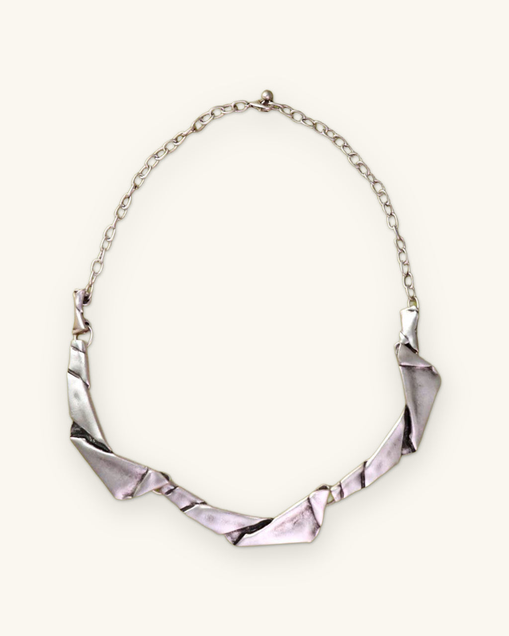 Folded-metal collar necklace with silver-tone link chain on an olive green backdrop, shown on a sand background