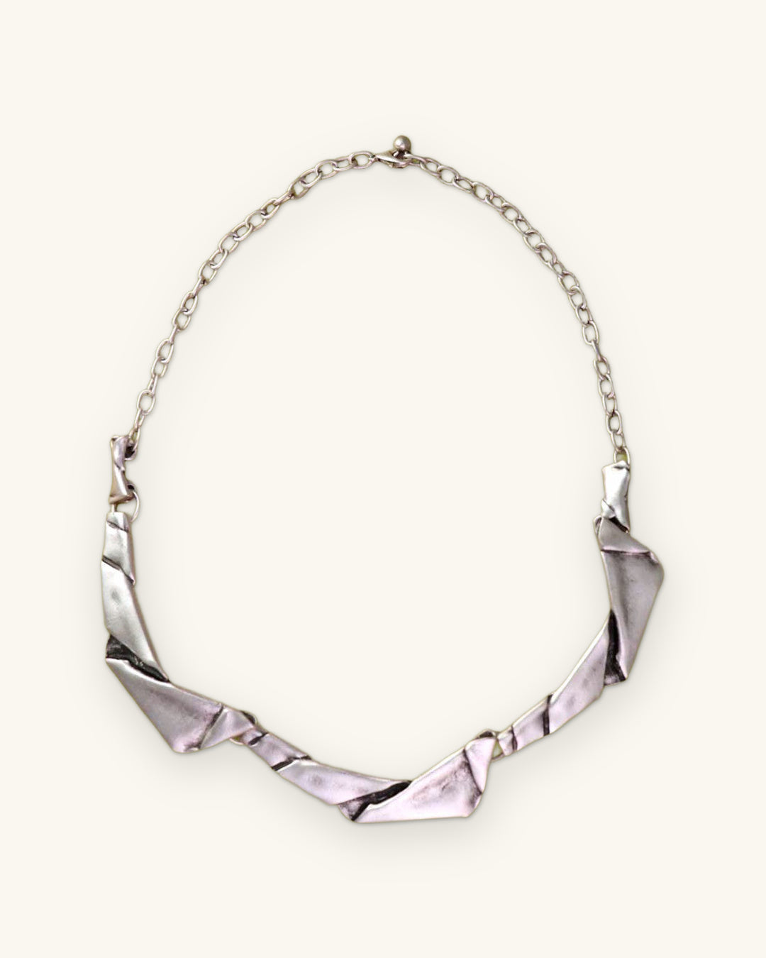 Folded-metal collar necklace with silver-tone link chain on an olive green backdrop, shown on a sand background