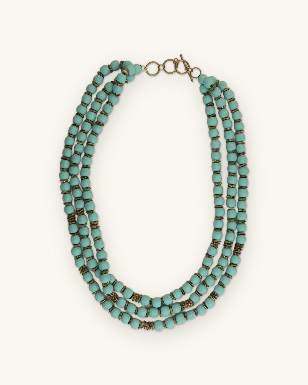 Powder blue triple-strand wood bead necklace with brass spacers and toggle closure, hand-turned in India, shown on a sand background
