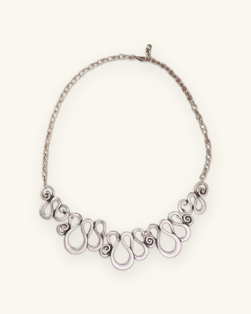 Scrollwork bib necklace with cable chain and lobster clasp laid on a blush surface, shown on a sand background
