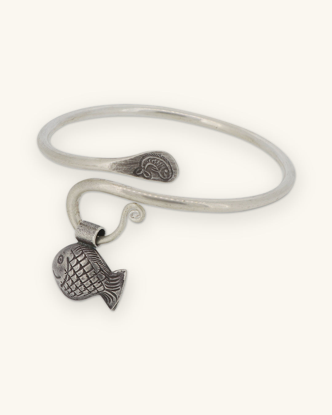 Silver open cuff bracelet with a textured fish charm, stamped spiral ends and curled wire loop, shown on a sand background