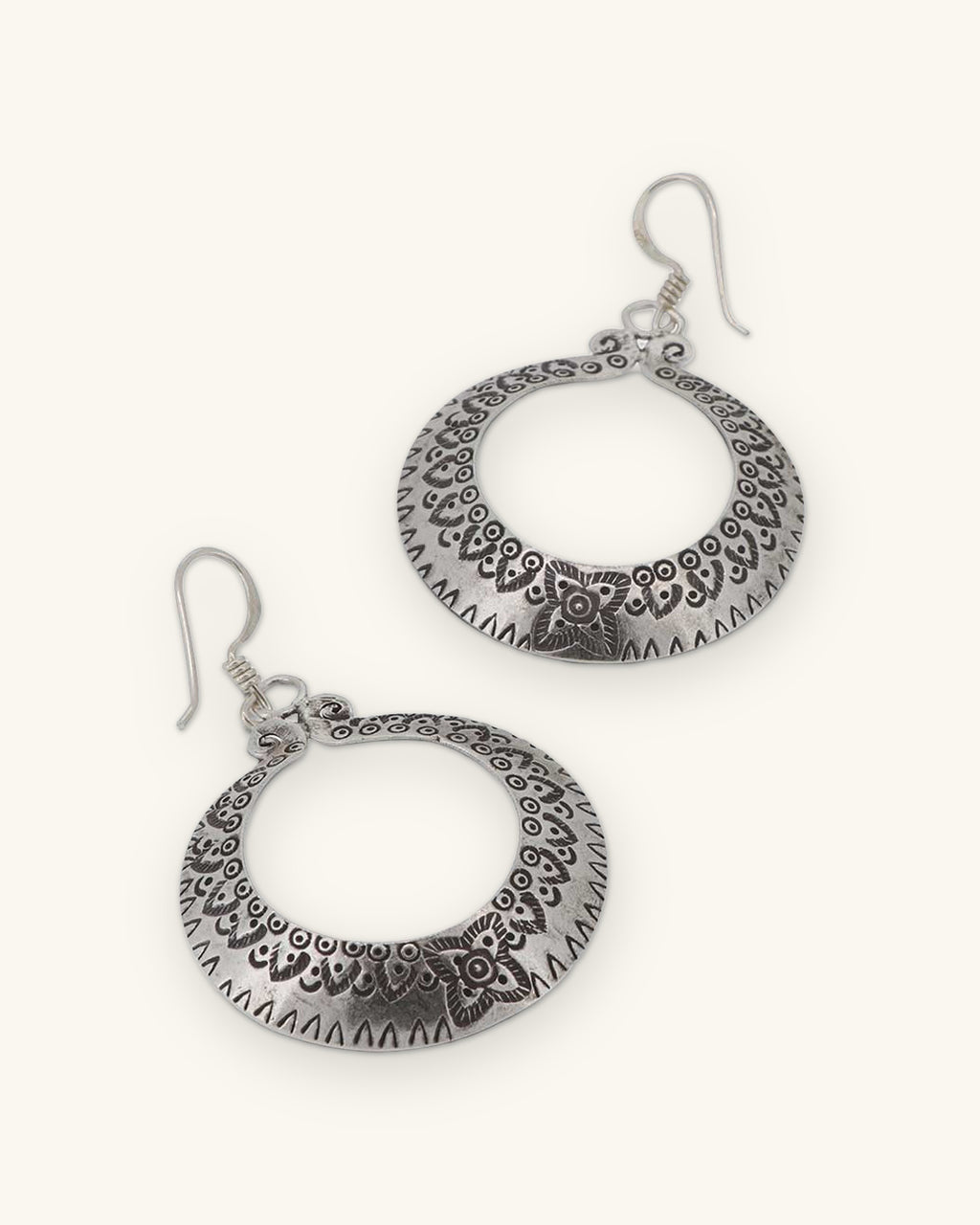 Pair of Thai Hilltribe silver crescent hoops with stamped floral border on French hook ear wires, shown on a sand background