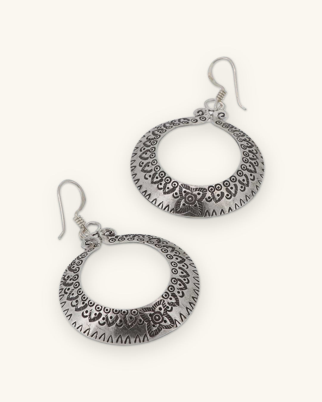 Pair of Thai Hilltribe silver crescent hoops with stamped floral border on French hook ear wires, shown on a sand background