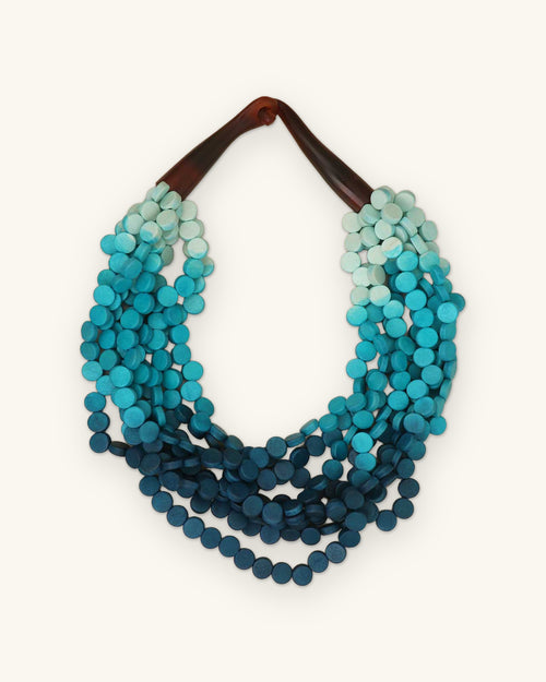 Aqua ombre wooden disc multi-strand necklace fading from pale aqua to deep teal, made in the Philippines, shown on a sand background